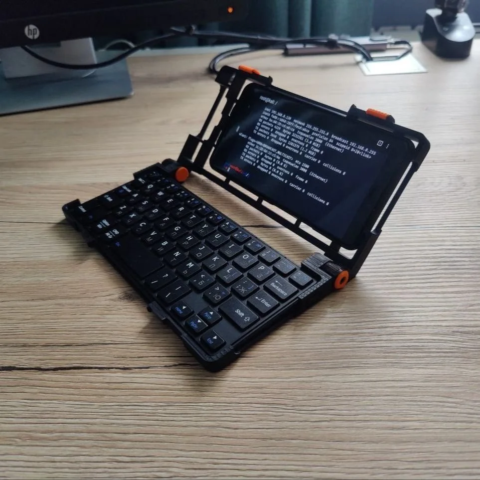 Un cyberdeck fai-da-te su una scrivania di legno: telaio nero con fermi arancioni, uno smartphone come schermo che mostra l'output di un terminale e una tastiera meccanica compatta